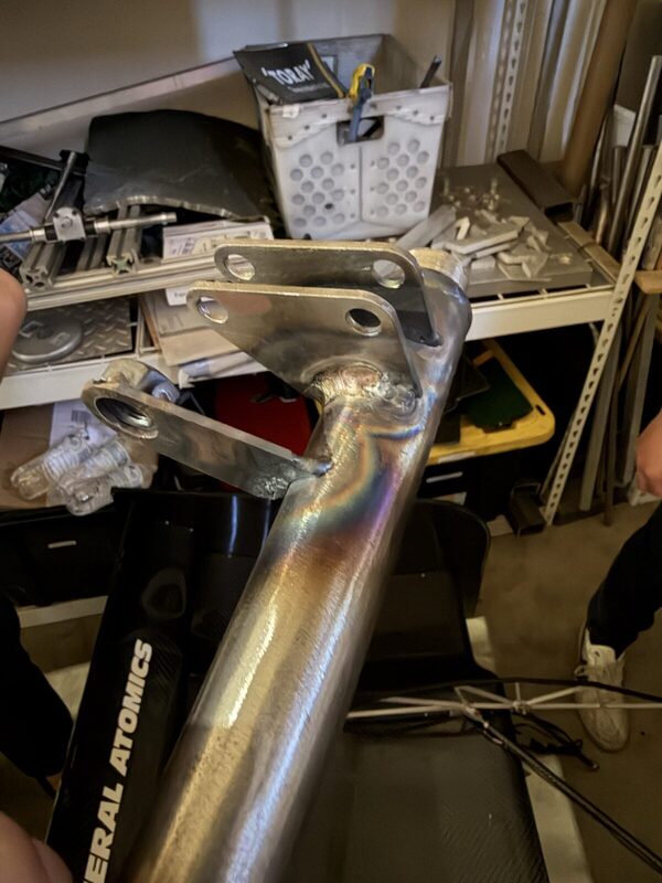 A freshly welded steel bracket with heat-tinted weld beads on a chassis tube, held up in the shop.